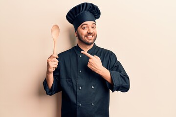 Young hispanic man wearing cooker uniform holding spoon smiling happy pointing with hand and finger