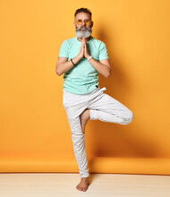 Portrait Of A Calm Modern Stylish Adult Man In Casual Style, Standing On One Leg And Holding Palms Together. Man Doing Yoga Exercises On An Orange Background. Concept Of Peace And Balance.