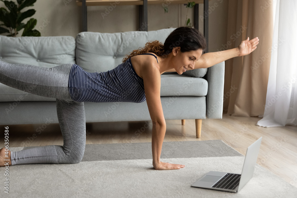 Sporty millennial Caucasian woman watch online sports lesson on laptop doing morning gymnastics in living room at home. Active young female train practice yoga or stretching on webcam on computer. - obrazy, fototapety, plakaty 