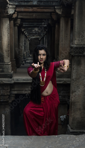 Portrait of beautiful Indian girl in heritage stepwell wearing traditional Indian red saree, gold jewellery and bangles holding religious plate. Maa Durga agomoni shoot concept