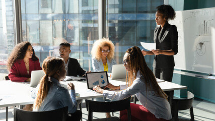 African American businesswoman lead meeting with multiethnic colleagues in office, discuss business ideas together. Ethnic male coach present project on flip chart at team briefing. Teamwork concept.