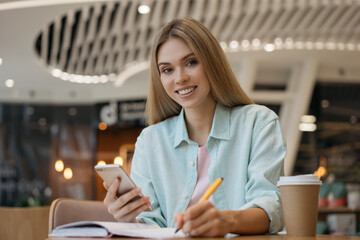 Young smiling woman freelancer  using mobile phone, taking tones, working in cafe. Portrait of happy student studying, exam preparation, education concept 