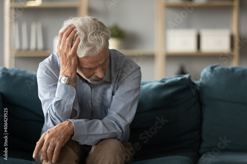 Frustrated unhappy middle aged mature man sitting on sofa, feeling depressed alone at home. Confused senior retired grandfather worrying about difficult life decision, copy space, old people solitude.