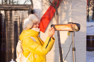 Portrait of a happy woman in a warm Russian hat on a frosty street warms her hands and a heater near a cafe and smiles broadly with a snow-white smile