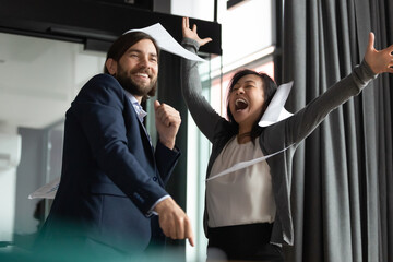 Poster - Overjoyed young 30s businessman in formal wear having fun with happy emotional middle aged korean asian female colleague, throwing papers in air, dancing celebrating corporate achievement in office.