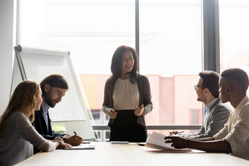 Poster - Happy mature korean asian female leader holding negotiations briefing meeting with motivated employees in modern office. Smiling skilled mixed race businesswoman mentoring colleagues near flipchart.