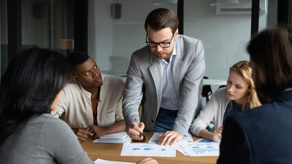 Poster - Skilled focused young businessman boss leader in eyeglasses analyzing sales data statistics or explaining marketing strategy to motivated diverse multiracial colleagues at brainstorming meeting.