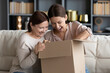 © fizkes - Overjoyed young woman with mature mother unpacking parcel together, looking into cardboard box, sitting on couch at home, happy elderly mum and grownup daughter excited by delivery, received order