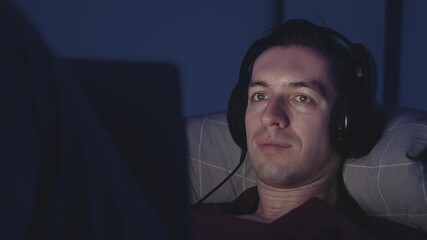 Young man in headphones watching film movie on laptop while resting on bed at night. Male using laptop computer while lying in the bedroom at home at nighttime.
