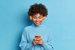 © wayhome.studio  - Smiling teenager with Afro hair listens favorite music track holds mobile phone downloads song to her playlist dressed in casual jumper isolated over blue background. People hobby technology