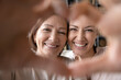 © fizkes - Close up happy smiling mature mother and grownup daughter taking selfie together, looking through fingers, making heart gesture with hands, family enjoying leisure time, having fun with camera,