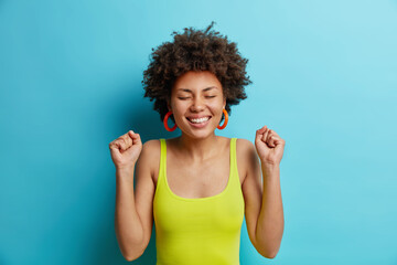 Wall Mural - Portrait of cheerful dark skinned young woman clenches fists with triumph happy to achieve goal celebrates winning competition closes eyes dressed in casual wear isolated over blue background.