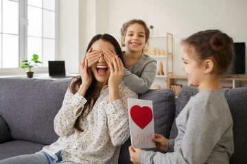 We love you, mommy. Excited young woman receiving present from kids. Little children wishing mother Happy Mother's Day. Two cute twin sisters cover mom's eyes as they give her handmade greeting card