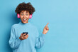 © WHstudio Leushin N - Glad ethnic woman with Afro hair indicates at upper right corner holds modern cellular listens music via stereo headphones dressed in loose knitted sweater isolated over blue background copy space