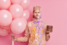 Unhappy Displeased European Man Frowns Face Holds Bunch Of Balloons Cake Has Spoiled Holiday Dressed In Festive Clothes Crown On Head Isolated Over Pink Background. People Party Celebration Concept