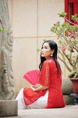 Poster - Pensive pretty young woman in traditional Vietnamese dress sitting outdoors with paper fan in hands