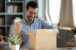 Happy 30s young man in eyewear looking inside of carton box, celebrating getting wished item or gift from internet store, having positive online shopping experience, fast postal delivery service.