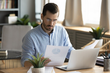 Concentrated young businessman ceo manager in eyewear looking at marketing research report, analyzing statistics data in charts, developing growth strategy, working on computer alone in modern office.