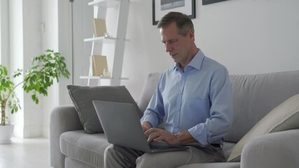 Smiling happy middle aged 50s old senior man using laptop computer technology working online, surfing internet, typing email, shopping in ecommerce store relaxing with device sitting on couch at home.