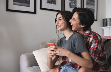 Wall Mural - Young happy lesbian couple hugging, laughing, holding smartphone at home. Cheerful funny girls best friends having fun using mobile apps together. Generation z people mobile technology users lifestyle