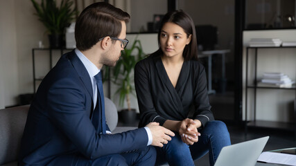 Wall Mural - Close up confident mentor wearing glasses training Indian intern, explaining task, sitting on couch in modern office with laptop, businessman and businesswoman working together, manager consulting