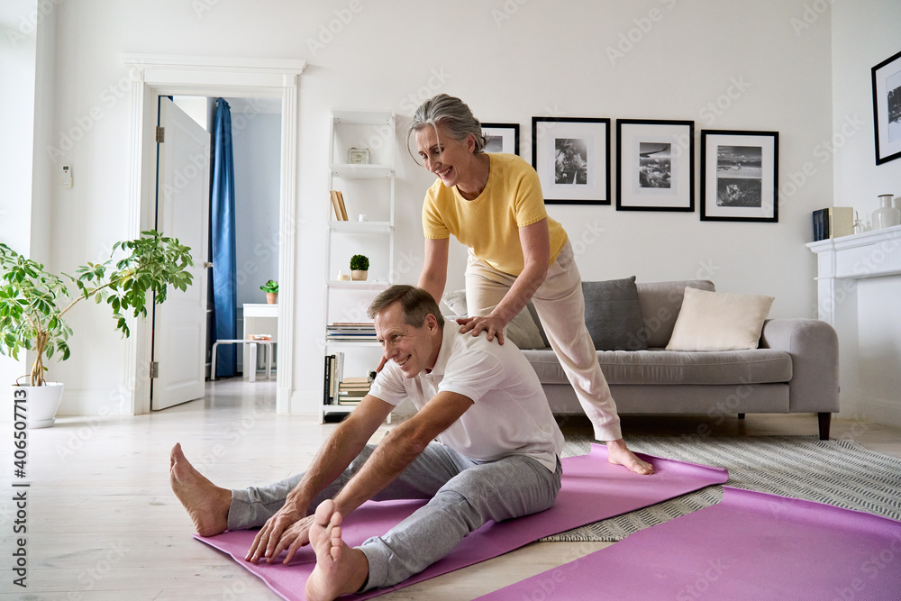 Fit active retired middle aged wife helping senior husband doing stretching exercise at home. Happy healthy older senior 60s couple enjoying fitness sport training workout together in apartment. - obrazy, fototapety, plakaty 