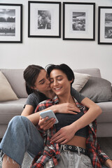 Wall Mural - Young happy generation z lesbian lgbtq diverse ethnicity couple hugging, holding smartphone, enjoying using mobile phone technology device apps, chilling at home siiting in living room together.