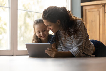 Caring young mom and little daughter lying on warm home floor have fun studying learning using modern pad gadget. Happy mother and small girl child look at tablet screen watching video online.