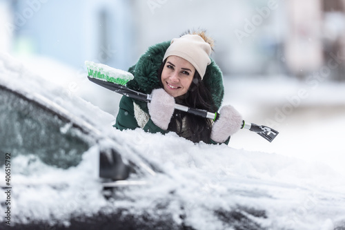Smiling well clothed woman holds ice scraper and snow broom while leaning against the car covered in snow