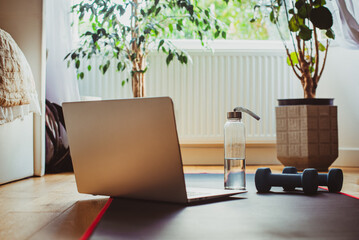 A glass water bottle, laptop, yoga mat and dumbbells on the floor in the bedroom for home exercising. Home online workout concept. Fitness online training. Soft selective focus. Copy space.