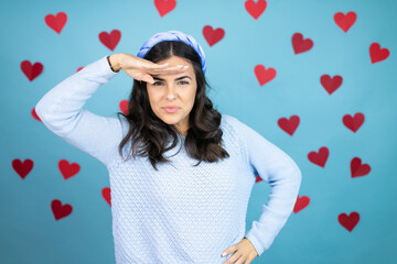 Young beautiful woman over blue background with red hearts very happy and smiling looking far away with hand over head. searching concept.