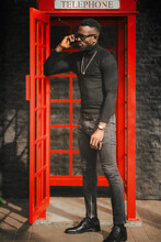 Portrait Of African Model Using Smartphone While Smoking Cigar In Telephone Booth