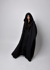 Wall Mural - Full length portrait of pretty black haired woman wearing long dark gown and a cloak.  Standing pose facing away from the camera, against a  studio background.