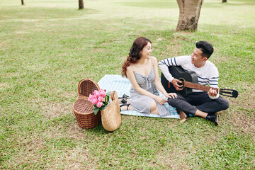 Wall Mural - Handsome young Asian man playing guitar and singing for his girlfriend when they are having picnic in park