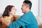 Fototapeta  - Happy young Asian boyfriend and girlfriend sitting on sofa, hugging and looking at each other