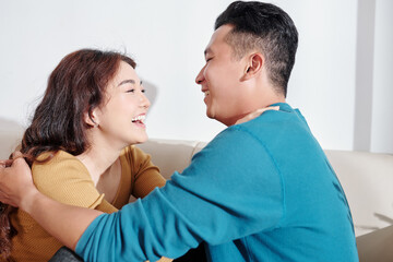 Wall Mural - Happy young Asian boyfriend and girlfriend sitting on sofa, hugging and looking at each other