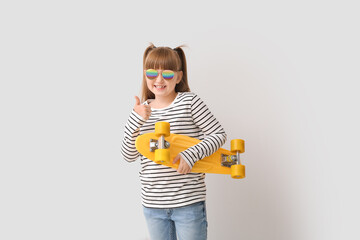 Poster - Cute little girl with stylish sunglasses and skateboard showing thumb-up gesture on light background