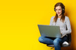 Young woman in headphones sitting on chair and holding laptop on isolated yellow background