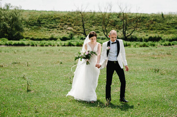 Wall Mural - Bride and groom walking and holding bouquet of flowers and greens, greenery on nature. Romantic couple newlyweds outdoors. Wedding ceremony.