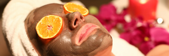 Wall Mural - Woman undergo wellness course and face cleansing. Client lye on massage table with his hair wrapped in towel, coal mask on her face, and orange circles in front of her eyes.