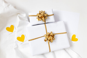 Two gift boxes in white wrapping paper on white table and on white textile with yellow paper hearts and white postcard. Wrapping paper. Mockup. Valentines day concept. Gold ribbon. 