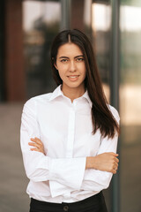 Portrait of a Caucasian pretty business lady smiling to the camera and crossing her hands while standing on the glass building background outdors