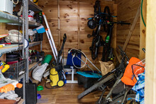 Suburban Home Wooden Storage Utility Unit Shed With Miscellaneous Stuff On Shelves, Bikes, Exercise Machine, Ladder, Garden Tools And Equipment. Messy And Chaos At House Yard Barn. Organization Order