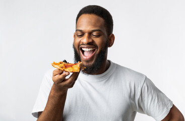 Wall Mural - Joyful African Millennial Guy Eating Pizza Slice Over White Background