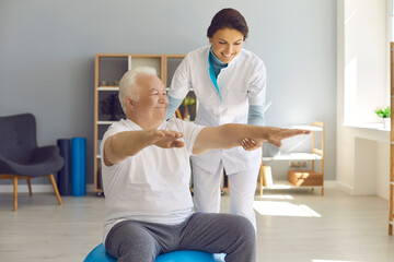 Happy senior man doing medical exercise on fitball with friendly nurse helping him