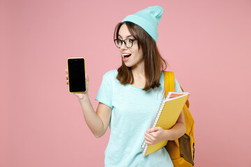 Wall Mural - Excited woman student in blue t-shirt hat glasses backpack hold notebooks mobile cell phone with blank empty screen isolated on pink background. Education in high school university college concept.