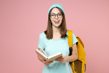 Wall Mural - Cheerful young woman student in casual blue t-shirt hat glasses backpack reading book isolated on pastel pink color background studio portrait. Education in high school university college concept.