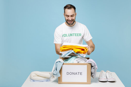 Funny young man in white volunteer t-shirt standing near table packing clothes in donation box for needy isolated on blue background studio. Voluntary free work assistance help charity grace concept.