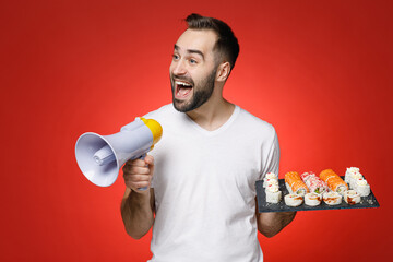 Poster - Excited young bearded man 20s wearing basic white t-shirt screaming in megaphone hold makizushi sushi roll served on black plate traditional japanese food isolated on red background studio portrait.