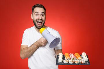 Poster - Surprised young bearded man 20s wearing basic white t-shirt screaming in megaphone hold makizushi sushi roll served on black plate traditional japanese food isolated on red background studio portrait.